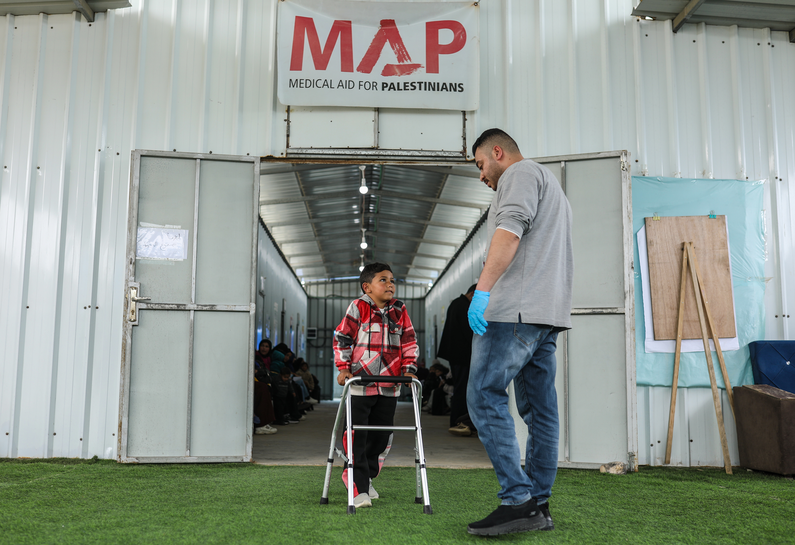 Yaqoub walking in MAP's Solidarity Polyclinic, supported by a walking aid and a MAP colleague's support.