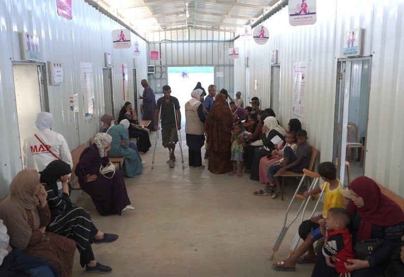 People sit on chairs lining a corridor in a clinic waiting for their appointments.