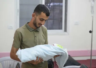A Palestinian father holds his baby swaddled in blankets, he looks down adoringly at the baby's face.