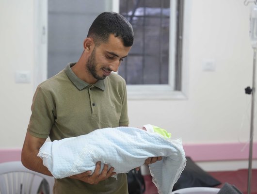 A Palestinian father holds his baby swaddled in blankets, he looks down adoringly at the baby's face.