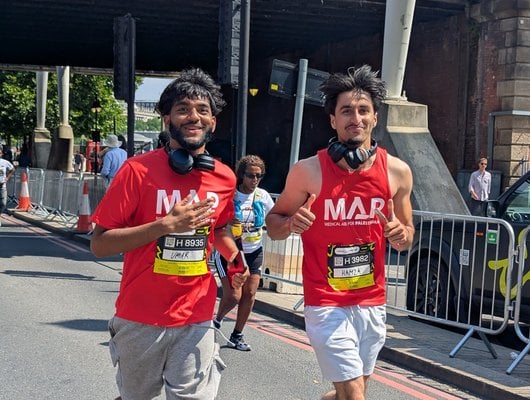 Two people run in the London 10k race. They are both wearing red MAP t shirts and are smiling at the camera as they run.