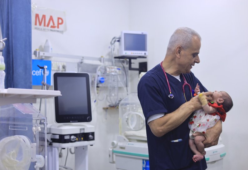 A small baby wearing a dress with cherries on it, in the arms of a doctor who is smiling down at her.