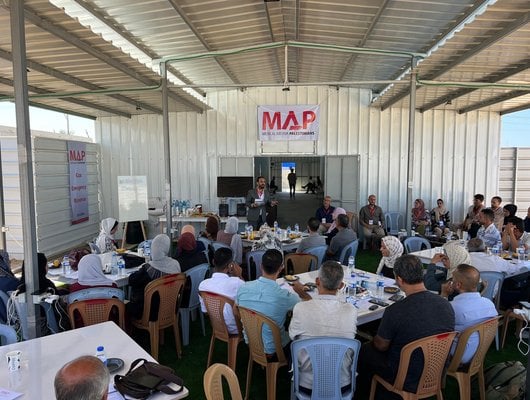 People sit around square tables at MAP's Solidarity Polyclinic in Gaza. A man stands in front of the tables delivering a talk.