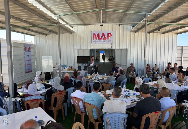 People sit around square tables at MAP's Solidarity Polyclinic in Gaza. A man stands in front of the tables delivering a talk.