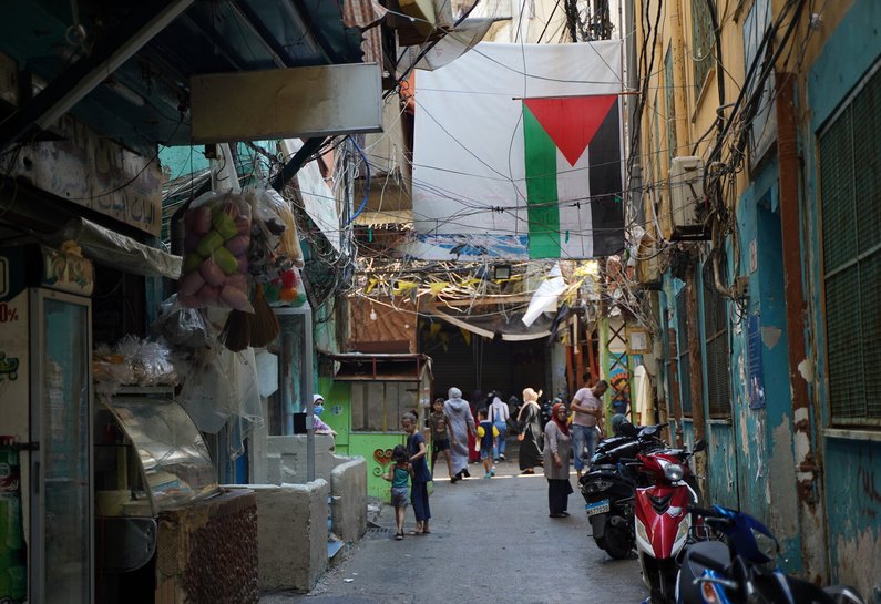 A Palestinian flag hangs over a street. Scooters are parked on one side of the street and a small shop opens onto the street on the other side. People are walking up and down the street.