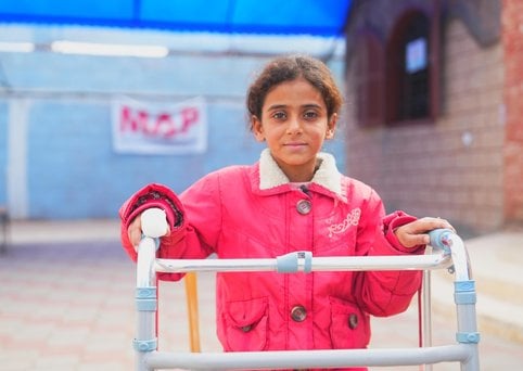 A girl wearing a red winter coat stands facing the camera supported by a metal walking frame.