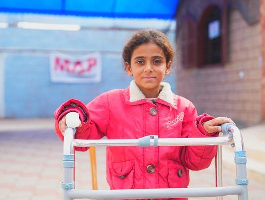 A girl wearing a red winter coat stands facing the camera supported by a metal walking frame.
