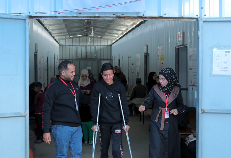 Maher walking out of the MAP's solidarity polyclinic in Gaza on crutches, accompanied by two MAP health care workers, all smiling.