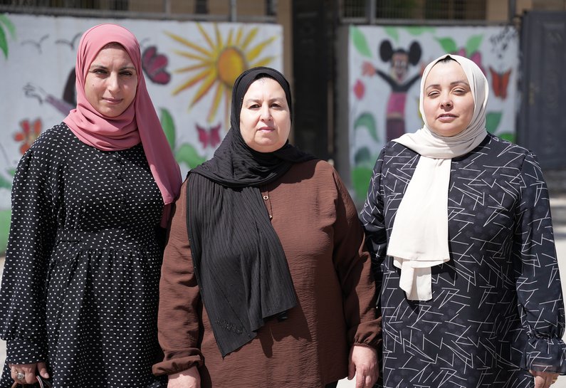 Three Palestinian women wearing hijabs look directly at the camera. The sun shines brightly on their faces.