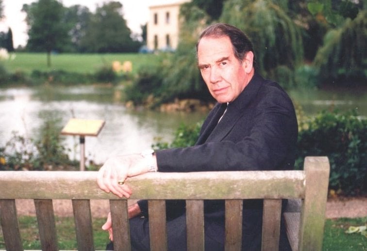 A man seated on a bench in front of a lake. He looks over the back of the bench directly at the camera.
